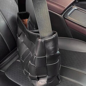 Puede incluir: Bolso de mano de cuero negro tejido con una correa para el hombro. El bolso tiene un dise&ntilde;o distintivo de tejido de cesta y una forma rectangular. El interior del bolso es visible, revelando un forro marr&oacute;n. El bolso est&aacute; sobre un asiento de coche de cuero negro.