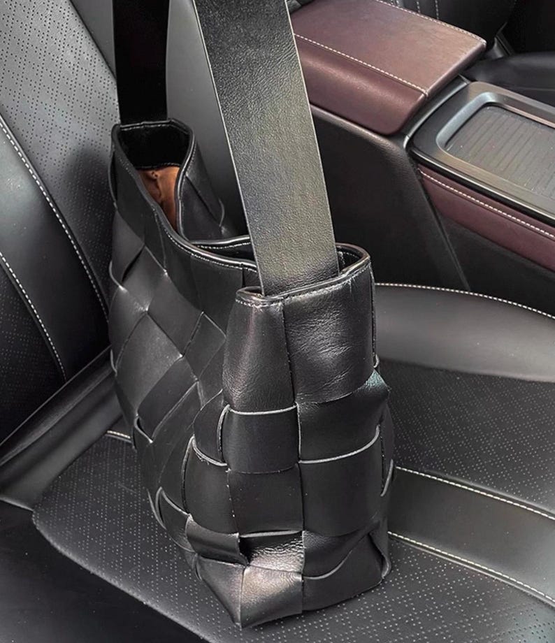 Puede incluir: Bolso de mano de cuero negro tejido con una correa para el hombro. El bolso tiene un dise&ntilde;o distintivo de tejido de cesta y una forma rectangular. El interior del bolso es visible, revelando un forro marr&oacute;n. El bolso est&aacute; sobre un asiento de coche de cuero negro.