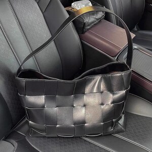 Puede incluir: Bolso tote de cuero negro tejido con una sola correa para el hombro. El bolso tiene forma rectangular con un patr&oacute;n tejido en la parte delantera y los lados. El interior del bolso es visible y parece ser de un color m&aacute;s oscuro. El bolso est&aacute; sobre un asiento de coche de cuero negro.