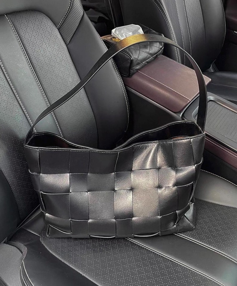 Puede incluir: Bolso tote de cuero negro tejido con una sola correa para el hombro. El bolso tiene forma rectangular con un patr&oacute;n tejido en la parte delantera y los lados. El interior del bolso es visible y parece ser de un color m&aacute;s oscuro. El bolso est&aacute; sobre un asiento de coche de cuero negro.