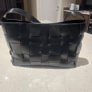 Puede incluir: Un bolso tote de cuero negro tejido con una sola correa para el hombro. El bolso presenta un dise&ntilde;o de tejido de cesta y una forma rectangular. El bolso est&aacute; sobre una superficie de color claro.