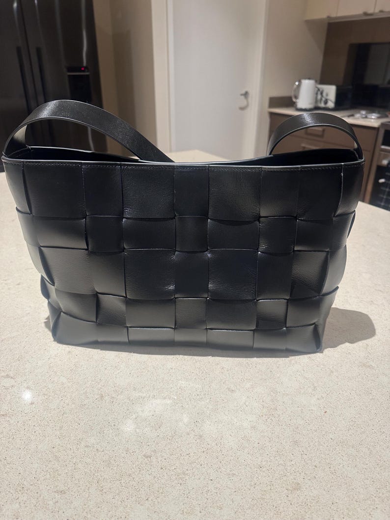 Puede incluir: Un bolso tote de cuero negro tejido con una sola correa para el hombro. El bolso presenta un dise&ntilde;o de tejido de cesta y una forma rectangular. El bolso est&aacute; sobre una superficie de color claro.