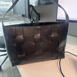 Puede incluir: Bolso tote de cuero negro con dise&ntilde;o tejido. El bolso tiene forma rectangular y una sola correa para el hombro. El interior del bolso es visible. El bolso est&aacute; sobre una superficie blanca.