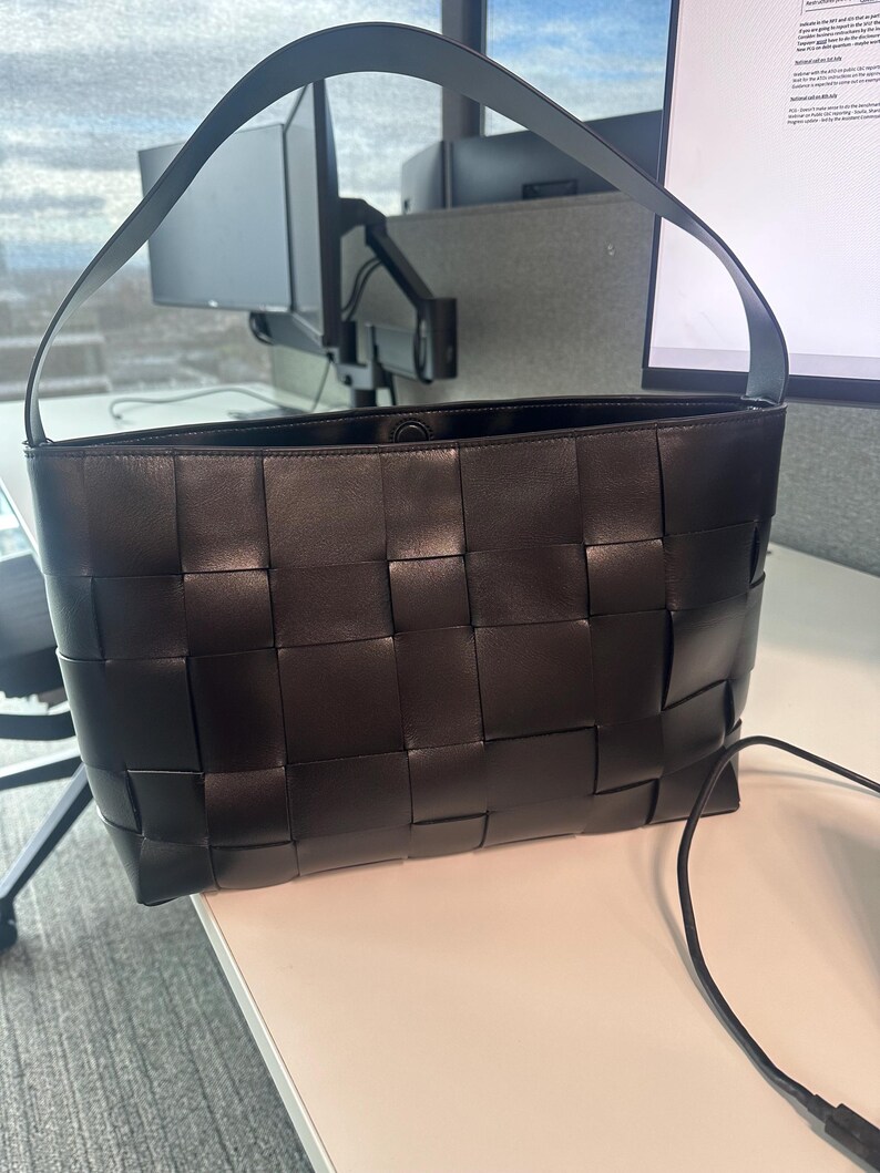 Puede incluir: Bolso tote de cuero negro con dise&ntilde;o tejido. El bolso tiene forma rectangular y una sola correa para el hombro. El interior del bolso es visible. El bolso est&aacute; sobre una superficie blanca.
