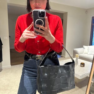 Puede incluir: Un bolso de hombro negro de cuero tejido, de forma rectangular y con una sola correa. El bolso es brillante. La persona de la imagen lleva una rebeca roja y vaqueros azul oscuro.
