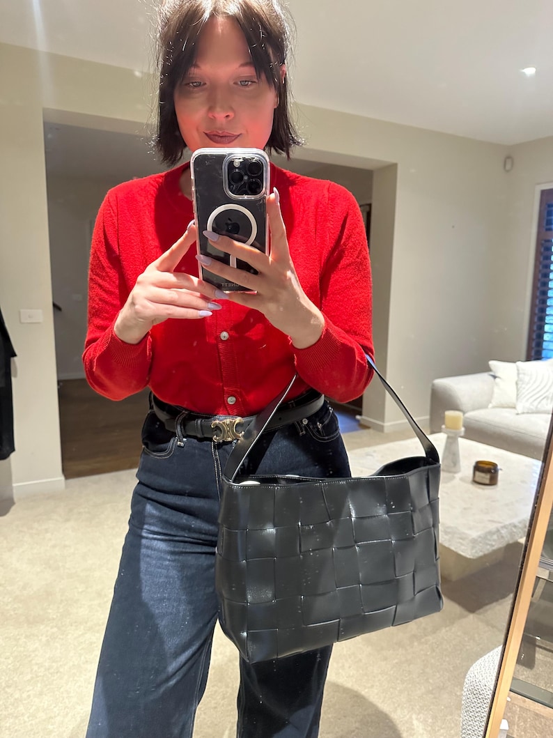 Puede incluir: Un bolso de hombro negro de cuero tejido, de forma rectangular y con una sola correa. El bolso es brillante. La persona de la imagen lleva una rebeca roja y vaqueros azul oscuro.