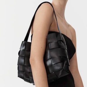 Puede incluir: Bolso tote de cuero negro tejido con correas largas para el hombro. El bolso tiene forma rectangular con un dise&ntilde;o tejido y se lleva al hombro. El bolso est&aacute; hecho de cuero liso y mate.