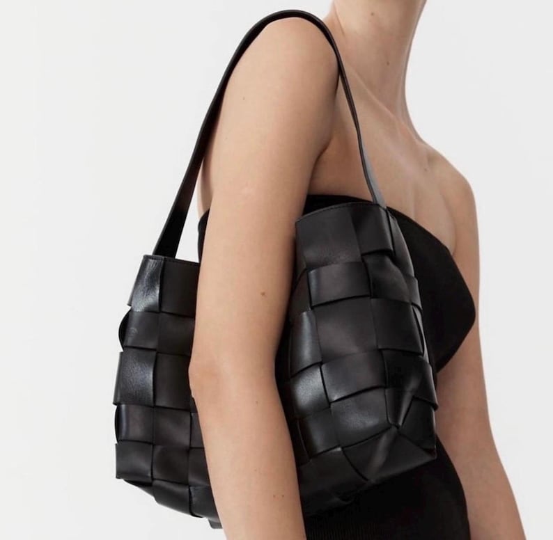 Puede incluir: Bolso tote de cuero negro tejido con correas largas para el hombro. El bolso tiene forma rectangular con un dise&ntilde;o tejido y se lleva al hombro. El bolso est&aacute; hecho de cuero liso y mate.