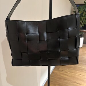 Puede incluir: Bolso de mano de cuero negro tejido con una sola correa para el hombro. El bolso tiene forma rectangular con un dise&ntilde;o de patr&oacute;n tejido. El bolso se exhibe en un soporte de metal negro.