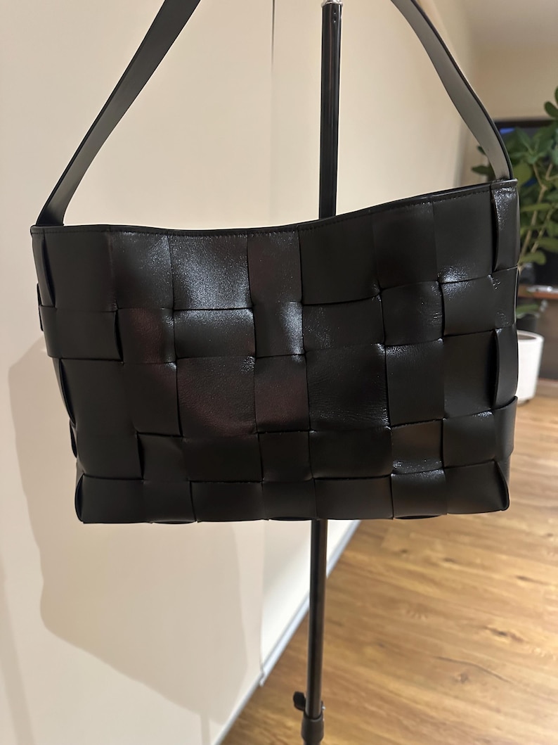 Puede incluir: Bolso de mano de cuero negro tejido con una sola correa para el hombro. El bolso tiene forma rectangular con un dise&ntilde;o de patr&oacute;n tejido. El bolso se exhibe en un soporte de metal negro.