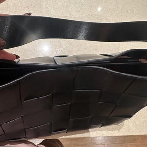 Puede incluir: Bolso de mano de cuero negro tejido con una correa ancha para el hombro. El bolso presenta un dise&ntilde;o distintivo de tejido de cesta. La correa es de color negro liso y el interior del bolso es visible. El bolso es de color oscuro y neutro.
