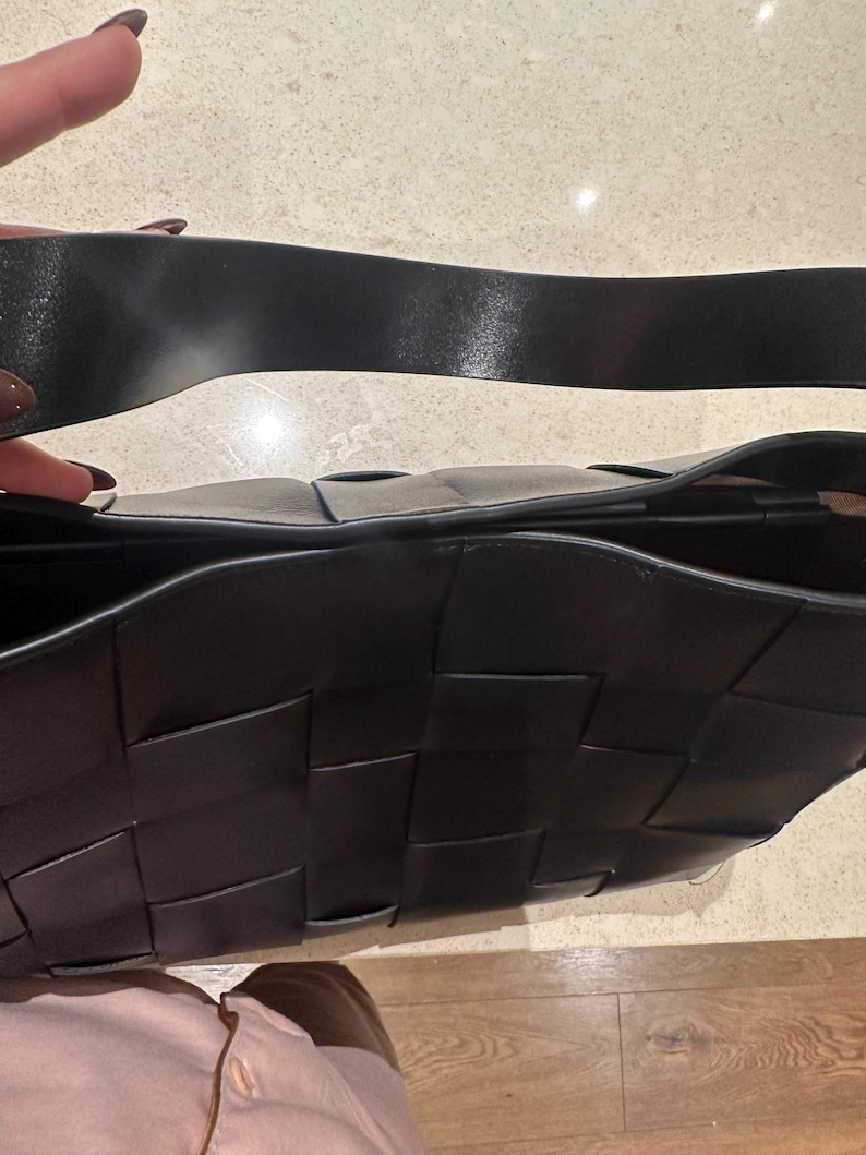 Puede incluir: Bolso de mano de cuero negro tejido con una correa ancha para el hombro. El bolso presenta un dise&ntilde;o distintivo de tejido de cesta. La correa es de color negro liso y el interior del bolso es visible. El bolso es de color oscuro y neutro.