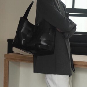 Puede incluir: Bolso tote de cuero negro con dise&ntilde;o tejido. El bolso tiene una correa larga para el hombro y se lleva sobre el hombro. El fondo es neutro.