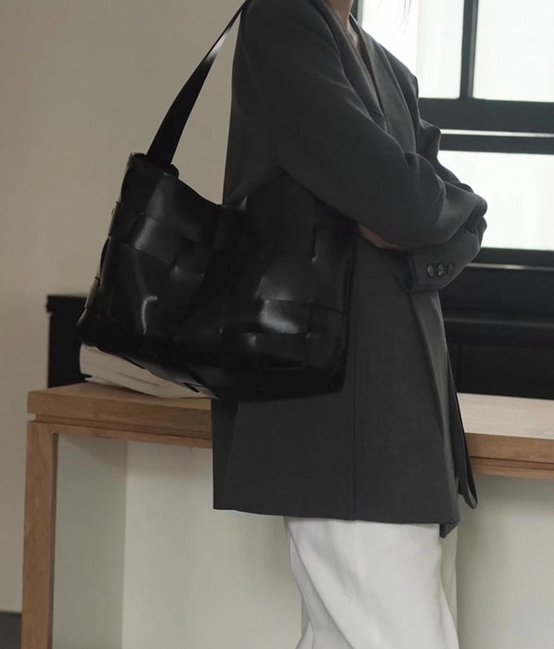 Puede incluir: Bolso tote de cuero negro con dise&ntilde;o tejido. El bolso tiene una correa larga para el hombro y se lleva sobre el hombro. El fondo es neutro.