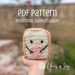 Puede incluir: Patrón de crochet para una hogaza de pan de apoyo emocional. La hogaza es blanca con un corazón rosa y ojos negros. El patrón está disponible para descargar en PDF. El texto "PDF Pattern emotional support Loadie" está en la parte superior de la imagen. El texto "Yarns of Joy" está en la parte inferior de la imagen.