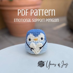 Könnte beinhalten: Ein gehäkelter Pinguin, der ein weißes Herz hält, mit dem Text "PDF Pattern Emotional Support Penguin" und "Yarns of Joy" auf dem Bild.