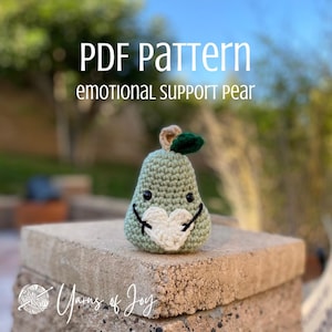May include: A handmade crocheted pear-shaped toy with a smiling face and a heart-shaped patch. The pear is light green with a brown stem and a green leaf. The image includes the text "PDF Pattern Emotional Support Pear" and "Yarns of Joy".