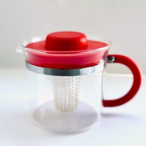 Könnte beinhalten: Eine rote Glas-Teekanne mit abnehmbarem Deckel und einem eingebauten Sieb. Die Teekanne hat einen klaren Glaskörper und einen roten Silikon-Griff.