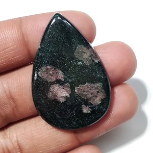 Op de afbeelding: Een druppelvormige, gepolijste edelsteen. De steen is voornamelijk donkergroen met onregelmatige vlekken van roze-paarse insluitsels. De edelsteen wordt in een hand gehouden tegen een witte achtergrond, waardoor de kleur en textuur worden benadrukt.