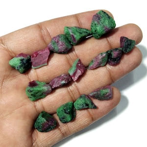 May include: A collection of rough-cut ruby in zoisite stones. The stones are a mix of deep red and vibrant green, with a raw, natural texture. The arrangement of the stones suggests they are for jewelry making or collecting.