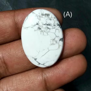 Pode incluir: Pedra preciosa oval branca com veios cinzentos. A pedra polida tem uma superfície lisa e um aspeto marmoreado. A pedra é segurada numa mão, e a letra "A" é visível no canto superior direito.