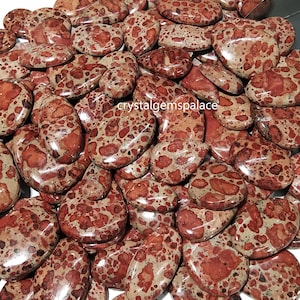 Könnte beinhalten: Eine Sammlung polierter, oval geformter Edelsteine. Die Steine weisen ein gesprenkeltes Muster aus rotbraunen Flecken auf beigem Hintergrund auf. Die Edelsteine sind glatt und glänzend, mit unterschiedlichen Größen und Formen. Das Bild ist gut beleuchtet.