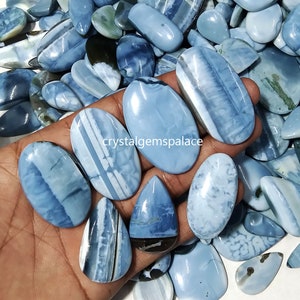 Pode incluir: Uma coleção de cabochões de opala azul em vários formatos e tamanhos. Os cabochões têm uma cor azul clara com bandas brancas e cinzas. Os cabochões são exibidos em uma mão.