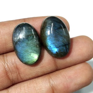 May include: Two oval labradorite gemstones. The stones show iridescent flashes of blue, green, and teal. The polished stones are held in a hand against a white background. The gemstones are approximately 2.5 cm in length.