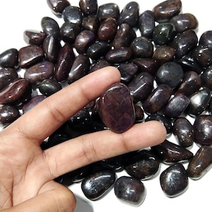 Può includere: Una collezione di pietre preziose lucide, rosso scuro-viola. Una pietra è tenuta tra le dita, mostrando la sua consistenza liscia e il colore. Le pietre preziose variano per dimensioni e forma, con una finitura lucida. Lo sfondo è bianco.