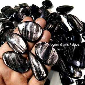 Peut inclure: Une collection de pierres précieuses noires polies avec des stries argentées iridescentes. Plusieurs pierres sont tenues dans une main, mettant en valeur leurs surfaces lisses et réfléchissantes. Les pierres précieuses sont de formes variées.