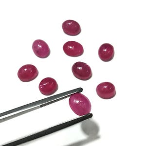 May include: A collection of oval-shaped, deep red gemstones. One gem is held by a pair of silver tweezers, showcasing its polished surface. The other gems are scattered on a white background, highlighting their vibrant color and smooth texture.
