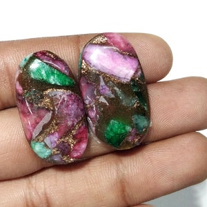 May include: Two oval-shaped gemstones with a mix of colors. The stones feature a blend of pink, green, and brown hues, with gold-colored veins running through them. The gemstones are held in a hand against a white background.