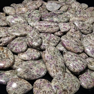 Pode incluir: Uma grande pilha de cabochões de pedra preciosa polidos em forma de lágrima, de cor verde e roxa. As pedras têm um padrão salpicado e veteado.