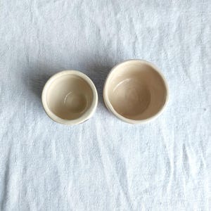 May include: Two white ceramic bowls, one smaller than the other, are shown from above on a white fabric background.