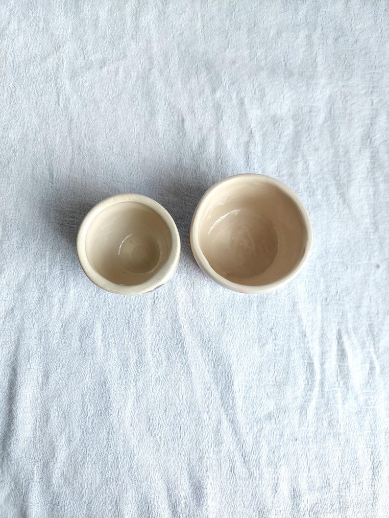 May include: Two white ceramic bowls, one smaller than the other, are shown from above on a white fabric background.