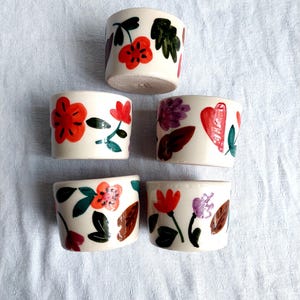 May include: Set of six small white ceramic cups with floral designs. Each cup features a different arrangement of red, orange, purple, and green flowers and leaves.