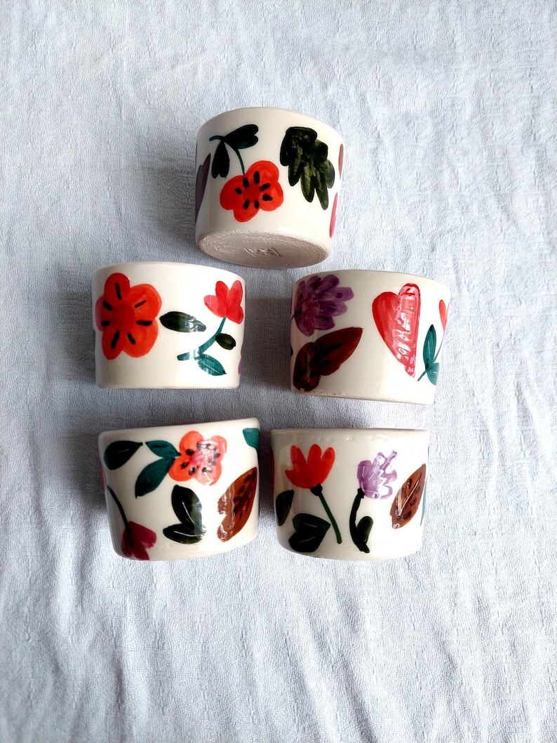 May include: Set of six small white ceramic cups with floral designs. Each cup features a different arrangement of red, orange, purple, and green flowers and leaves.