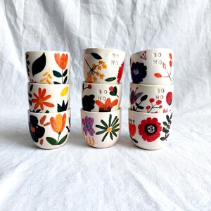 May include: A set of six small ceramic cups with floral designs. Each cup has a different floral pattern in shades of red, orange, yellow, green, blue, and purple. The cups are stacked in three groups of two. The cups are labeled "Bo Ho" in white lettering.