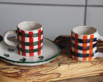 May include: Two ceramic coffee mugs with checkered patterns. One mug has a red and green checkered pattern, and the other has a blue and orange checkered pattern. Both mugs are on a wooden surface.