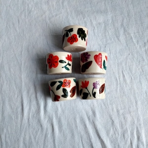 May include: Five small white ceramic cups with floral designs. The cups have red, orange, green, and purple flowers and leaves painted on them.