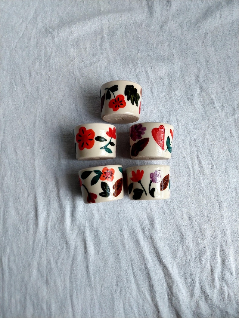 May include: Five small white ceramic cups with floral designs. The cups have red, orange, green, and purple flowers and leaves painted on them.
