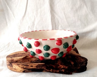 May include: White ceramic bowl with red and green polka dots. The bowl is sitting on a wooden stand.