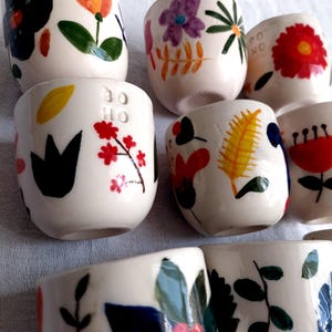 May include: Set of six small white ceramic cups with colorful floral designs. Each cup features a different floral pattern with various colors, including red, yellow, green, blue, and purple. The cups are hand-painted and have a unique, whimsical style.