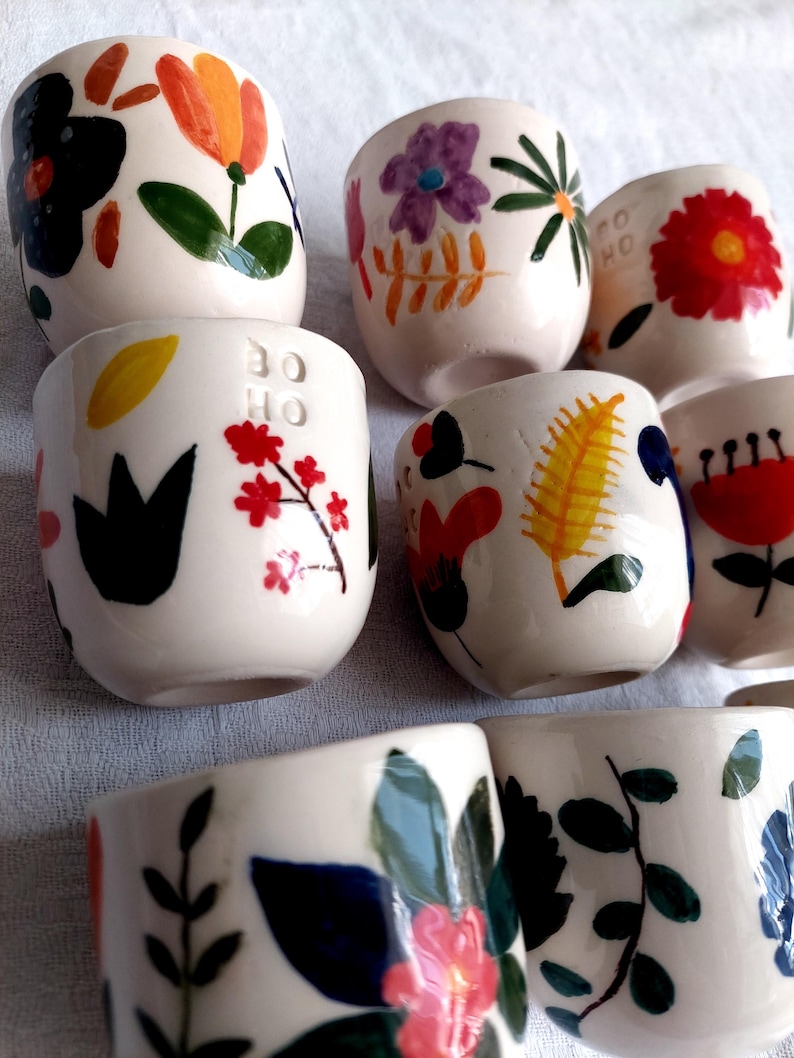 May include: Set of six small white ceramic cups with colorful floral designs. Each cup features a different floral pattern with various colors, including red, yellow, green, blue, and purple. The cups are hand-painted and have a unique, whimsical style.