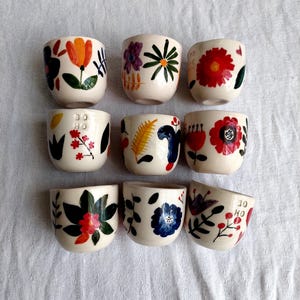 May include: Set of ten small white ceramic cups with colorful floral designs. Each cup features a different floral pattern with red, blue, yellow, green, and orange colors. Some cups have the text "DO HO" or "HO HO" written on them.