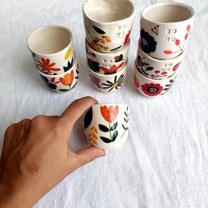 May include: A stack of six white ceramic mugs with floral designs in shades of orange, red, yellow, blue, and green. The mugs have the text "30 HO" written on them.