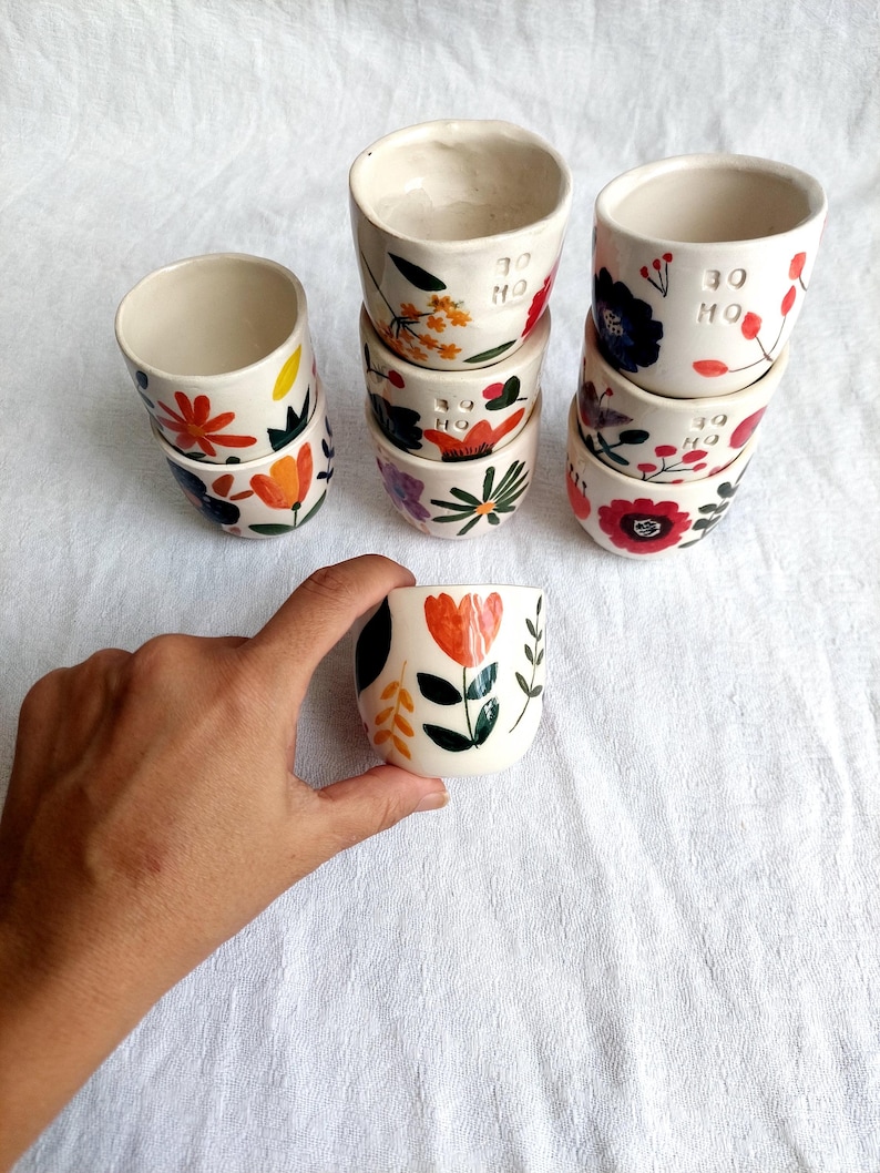 May include: A stack of six white ceramic mugs with floral designs in shades of orange, red, yellow, blue, and green. The mugs have the text "30 HO" written on them.