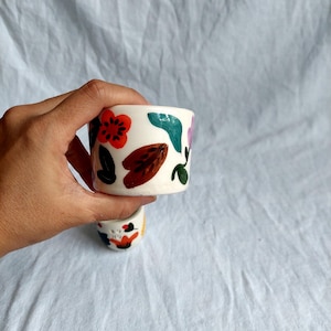 May include: Two white ceramic mugs with a floral design. The mugs are hand-painted with colorful flowers, leaves, and abstract shapes. The mugs are a unique and stylish addition to any kitchen or home.