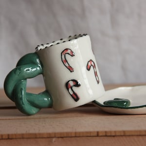 May include: A white ceramic mug with a green twisted handle and red and white candy cane designs. The mug is sitting on a white ceramic saucer with a green rim.