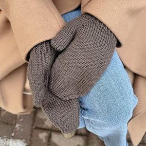 Puede incluir: Un par de mitones de punto marrón. Los mitones se sostienen juntos, con un abrigo color canela y unos vaqueros azules en el fondo. Los mitones parecen estar hechos de un hilo suave y cálido.